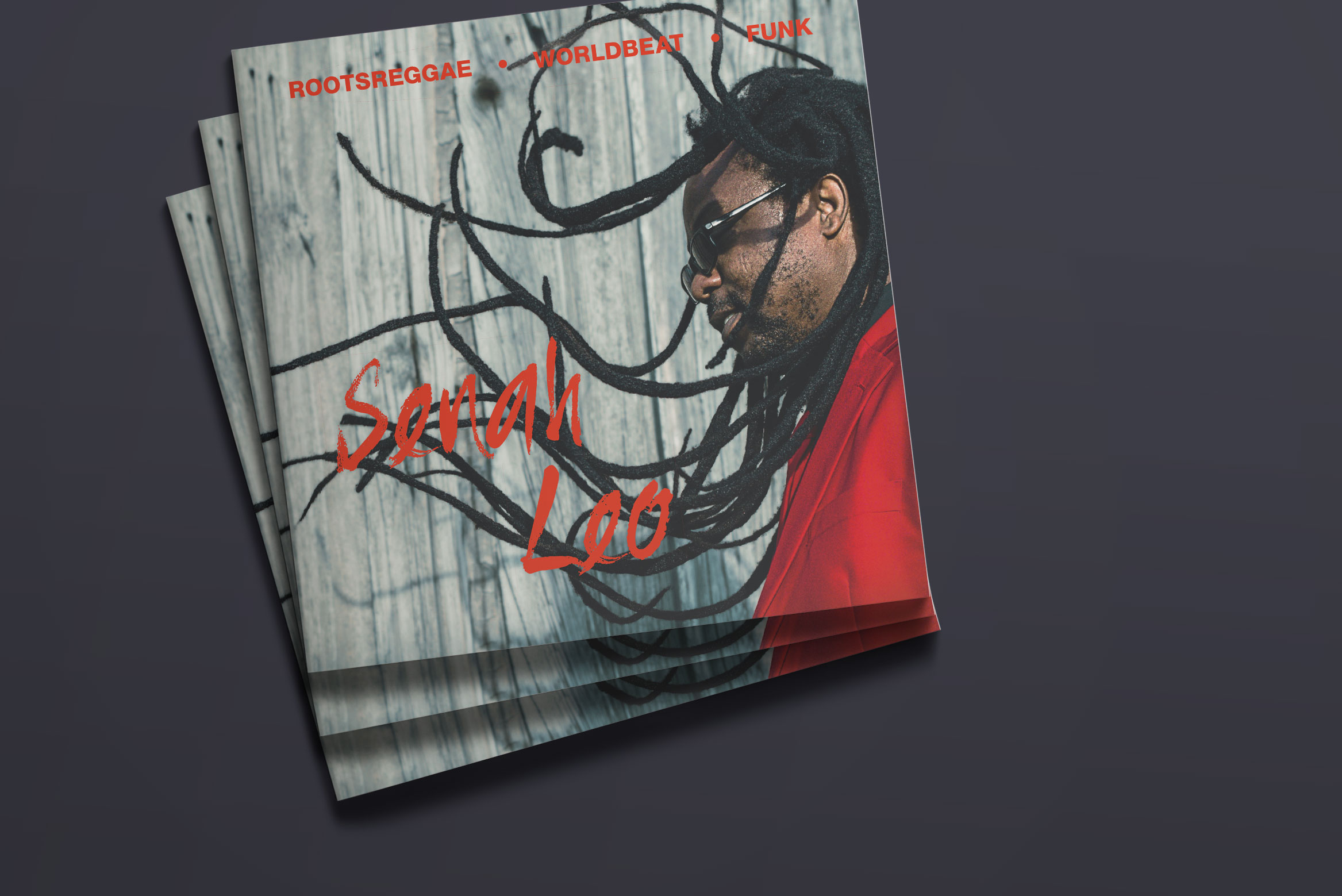 Der Header zeigt einen Stapel Flyer von Musiker Senah Leo aus Berlin auf dunklem Grund. Das Kommunikationsdesign zeigt den Musiker seitlich in rotem Anzug und dunkler Sonnenbrille und fliegenden Dreadlocks, darauf in rot die Unterschrift des Künstlers.