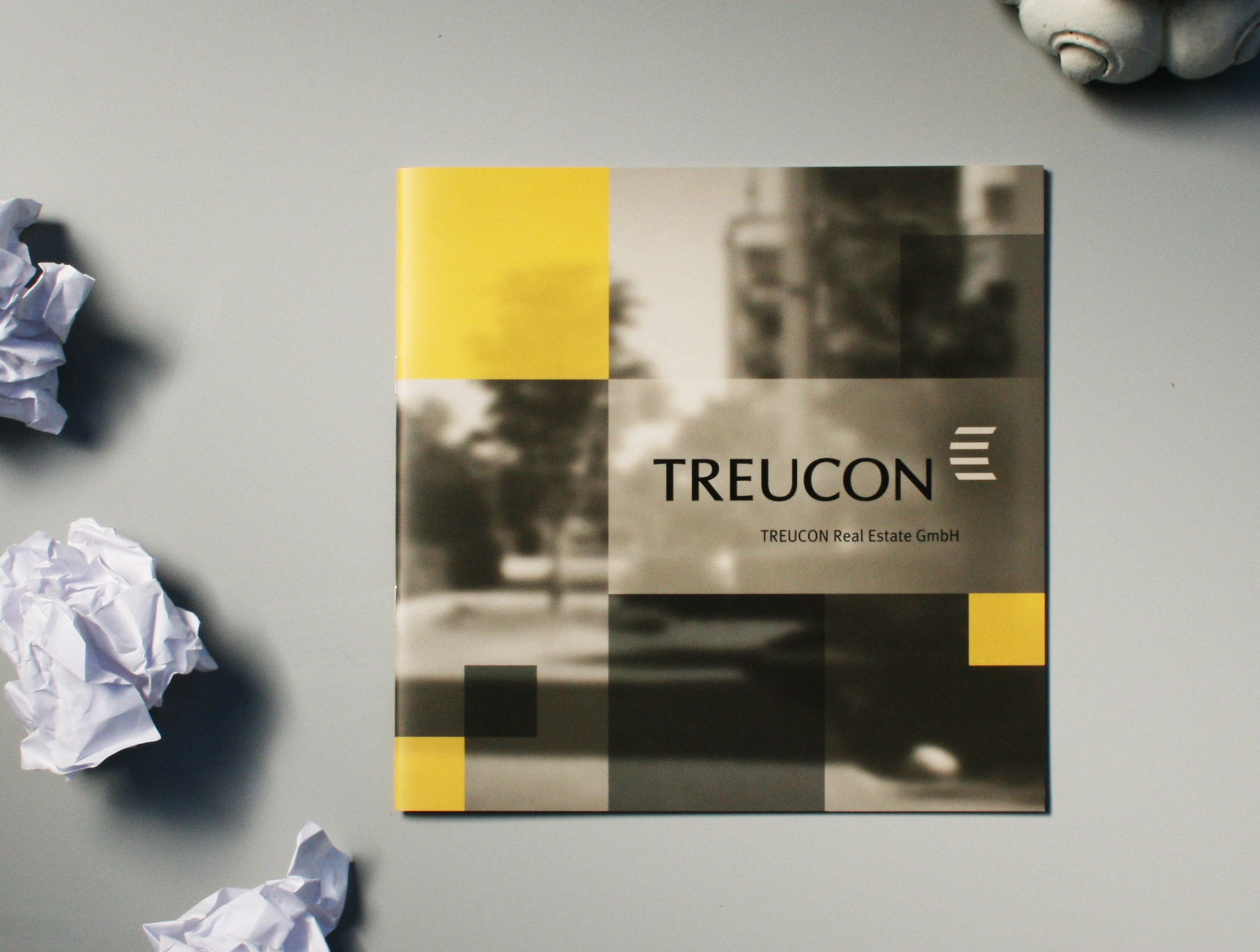 Mockup der Imagebroschüre Treucon Real Estate GmbH Berlin. Die Gestaltung des Covers besteht aus einer Collage mit tranzparenten schwarzen, gelben und weißen Flächen und einem diffusen Foto im Hintergrund, darauf ist das Logo von Treucon Real Estate platziert.