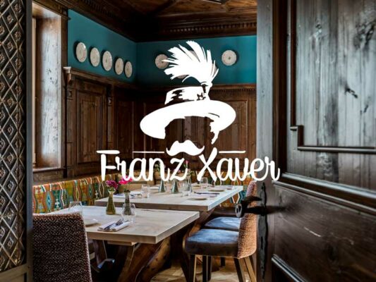 Das Logo vom Wirtshaus Franz Xaver wurde in weiß mittig auf einem Foto des Gaststätteninnenraums platziert.